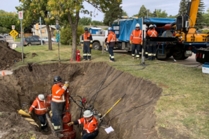 Photo of a crew wearing MWG FR Clothing. MWG FR Clothing is bright orange in colour with silver segmented reflective tape on torso and sleeves to meet high-visibility standard CSA Z96-15. Workers are seen wearing MWG Safety Apparel in hole working on a pipeline.