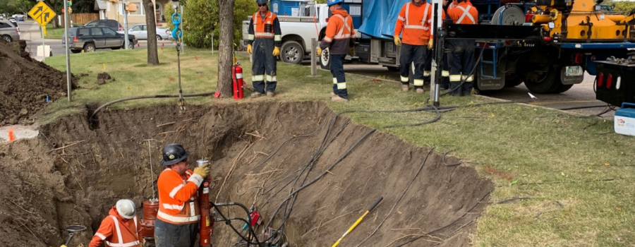 Photo of a crew wearing MWG FR Clothing. MWG FR Clothing is bright orange in colour with silver segmented reflective tape on torso and sleeves to meet high-visibility standard CSA Z96-15. Workers are seen wearing MWG Safety Apparel in hole working on a pipeline.