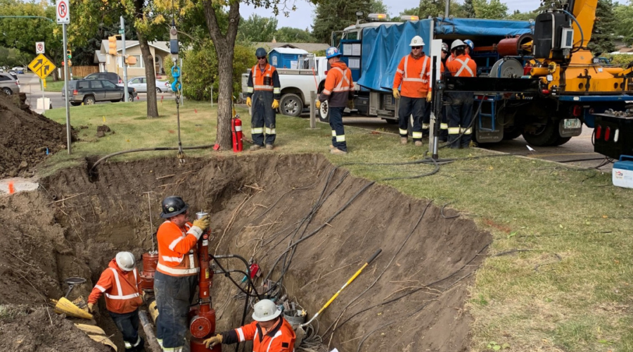 Photo of a crew wearing MWG FR Clothing. MWG FR Clothing is bright orange in colour with silver segmented reflective tape on torso and sleeves to meet high-visibility standard CSA Z96-15. Workers are seen wearing MWG Safety Apparel in hole working on a pipeline.